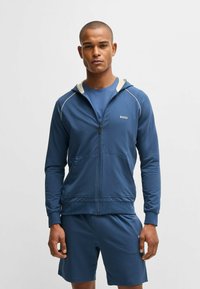 Blue zip-up hoodie with contrasting hood and white piping, worn over a matching blue t-shirt and shorts; logo on chest. Smooth texture.