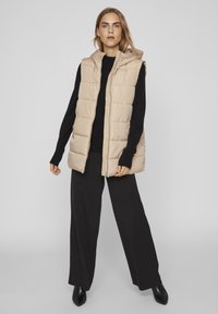 Gilet matelassé beige avec capuche, présentant un design matelassé et une fermeture éclair à l'avant. Porté par-dessus un pull noir et assorti à un pantalon large noir.
