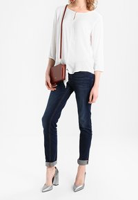 Blusa blanca de manga larga con un corte en forma de llave en la parte delantera, jeans ajustados oscuros y zapatos de tacón plateados. Completa con un bolso cruzado marrón.