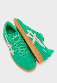 Rohelised sametist tenniseketid roosade detailidega, varustatud kummist tallaga, paeltega kinnitusega ja ventilatsiooni tagamiseks perforeeritud varvasega. Nähtav "Asics" brändimine.