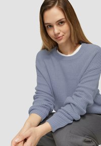 Hellblau strukturierter Strickpullover mit Rundhalsausschnitt und gerippten Bündchen, getragen über einem weißen Hemd. Model sitzt in einer lässigen Pose.