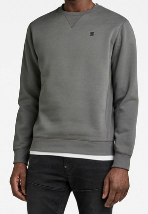 Homme portant un sweat-shirt gris uni à col rond avec un petit logo sur la poitrine gauche, associé à un pantalon noir et un sous-t-shirt blanc.