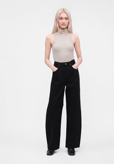 Jeune femme aux longs cheveux blonds portant un col roulé sans manches beige rayé et un pantalon noir à jambes larges, les mains dans les poches.