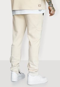 Beige Sweatpants mit schmaler Passform, gerippten Bündchen und seitlichen Taschen, kombiniert mit weißen Sneakers mit schwarzen Akzenten und Logo.