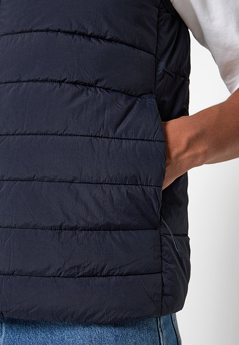 Chaleco acolchado azul marino con costuras horizontales, material ligero, bolsillos laterales y detalle de cremallera en el dobladillo. Lleva puesto sobre una camisa blanca.