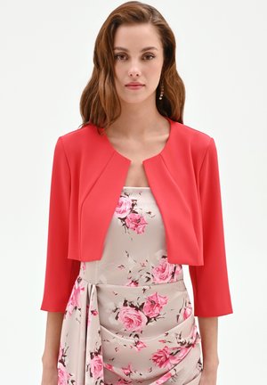 Donna con capelli lunghi castani che indossa una giacca corta rossa sopra un vestito beige chiaro con motivi floreali rosa e maniche a tre quarti.