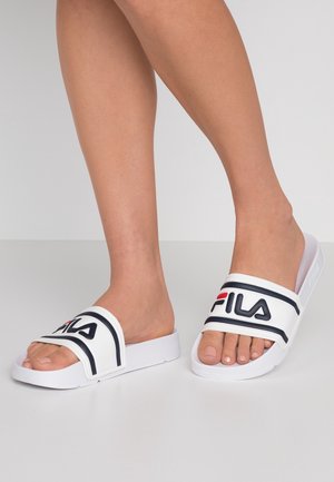 Ben iført hvide FILA slid-sandaler med sort og rød logo, stående på en lysegrå gulv mod en ensfarvet baggrund.
