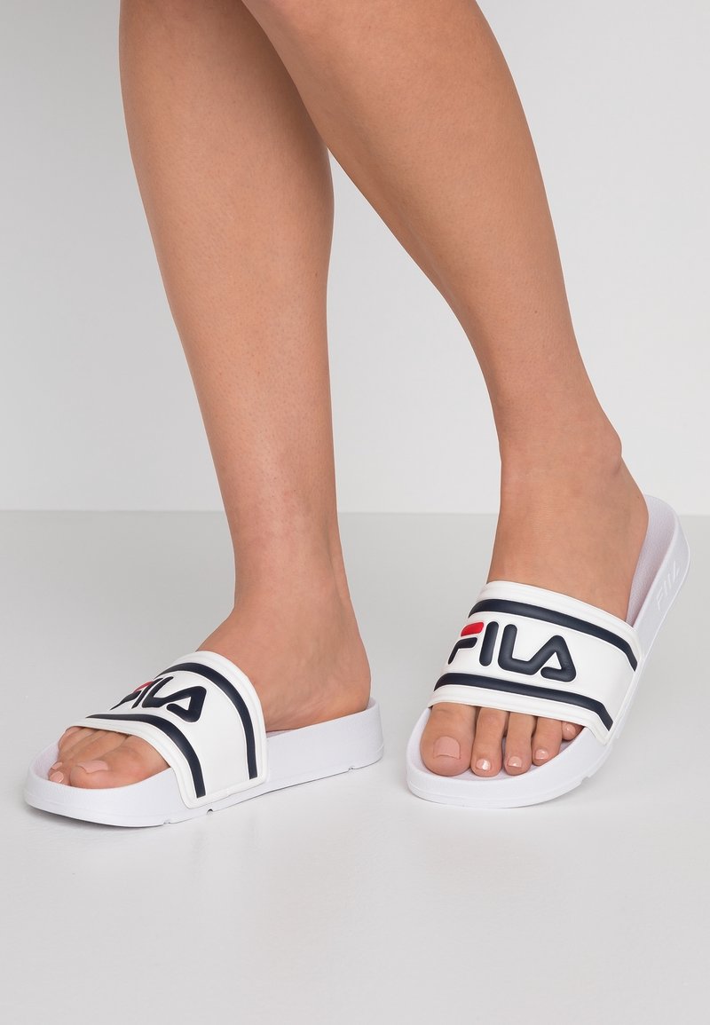 Jambes portant des sandales slides blanches FILA avec un logo noir et rouge, debout sur un sol gris clair sur un fond uni.