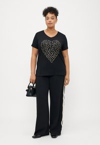 Μαύρη βαμβακερή μπλούζα με σχέδιο χρυσής μεταλλικής καρδιάς, συνδυασμένη με μαύρες φαρδιές παντελόνες και μαύρη τσάντα.