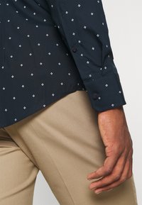 Marineblaues Hemd mit kleinen weißen Diamantmustern kombiniert mit beigen Hosen, zeigt Manschetten- und Seitennahtdetails am Arm und der Hand einer Person.