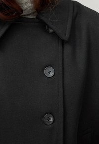 Manteau noir en tissu texturé, avec un col, quatre boutons noirs et des fermetures dissimulées sur le devant.