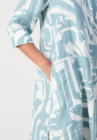 Bras d'une personne portant une chemise à manches longues bleue et blanche à motifs, avec la main placée dans une poche latérale.