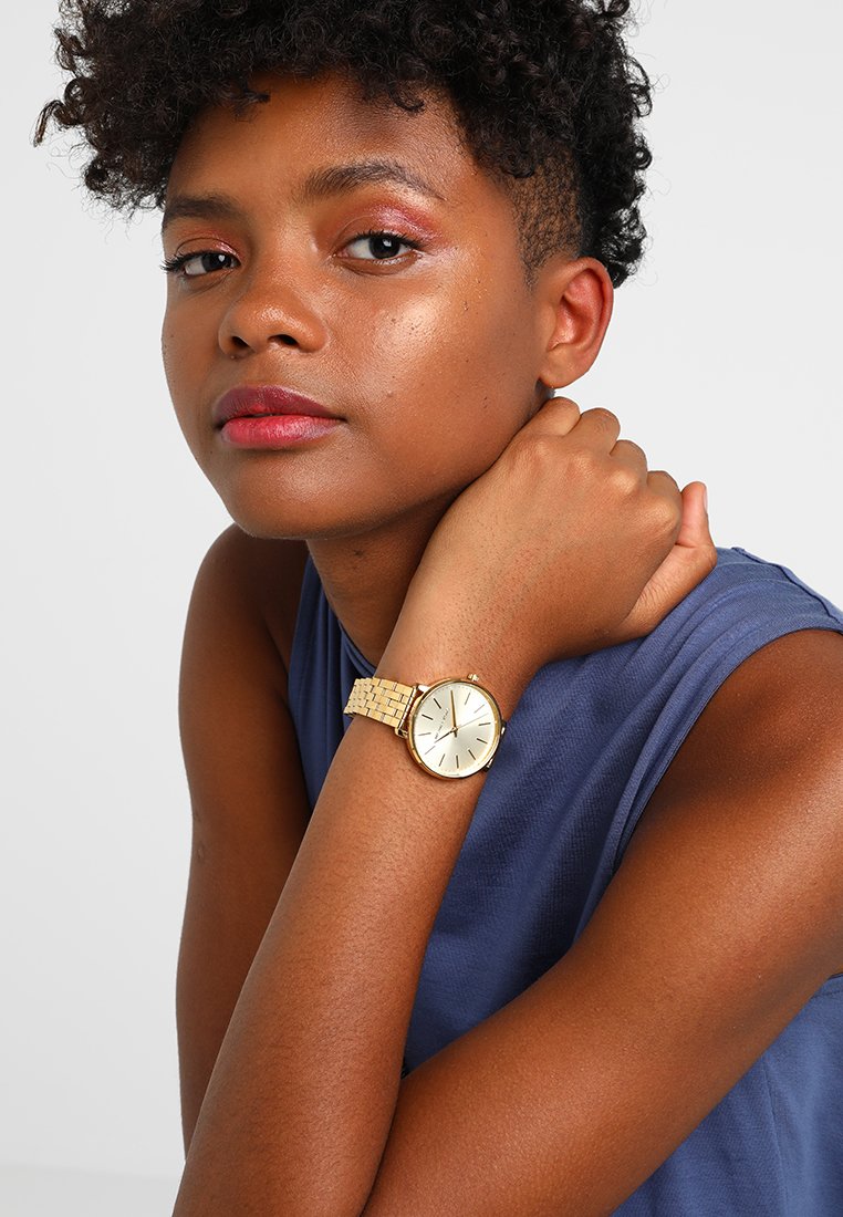 Gouden horloge met een textuur en patroon op de band. Ronde wijzerplaat met een witte achtergrond en slanke uurmarkeringen. Gedragen op een pols met een gebruinde huid.