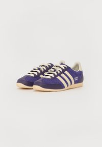 Baskets violettes en daim et cuir, avec des rayures crème, des détails perforés et une semelle en caoutchouc beige. Marquage sur le côté.