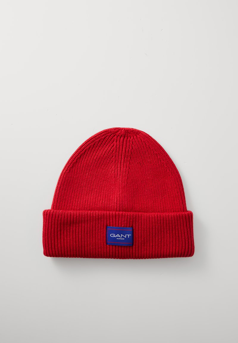 Red knit beanie with a ribbed texture and a folded brim. Features a blue rectangular tag with the brand name "GANT" in white letters.