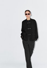 Vrouw draagt een zwart oversized jack, losse zwarte broek en donkere zonnebril, staand tegen een effen lichtgrijze achtergrond.