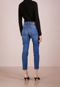 Vaqueros ajustados azules con cintura alta y bolsillos traseros, combinados con una blusa negra de manga larga y mules negros de tacón.