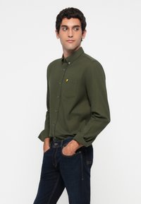 Hombre con cabello rizado oscuro que lleva una camisa de botones verde olivo y jeans oscuros, de pie con las manos en los bolsillos, mirando de lado.