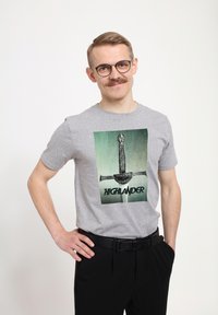 Šedé bavlněné tričko s grafikou meče a slovem "HIGHLANDER" v tučném černém písmu, krátké rukávy, nošené s černými kalhotami.