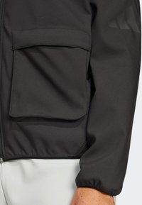 Black jacket featuring a zip front, elastic cuffs, and a large flap pocket. Smooth fabric with a matte finish and subtle logo detail on the sleeve.