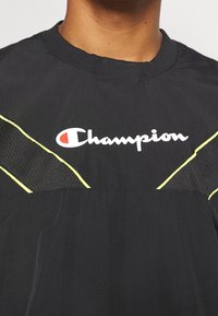 Czarna sportowa koszulka z siateczkowymi panelami i żółtymi wykończeniami. Na froncie widoczny duży biały logo "Champion". Lekki materiał.