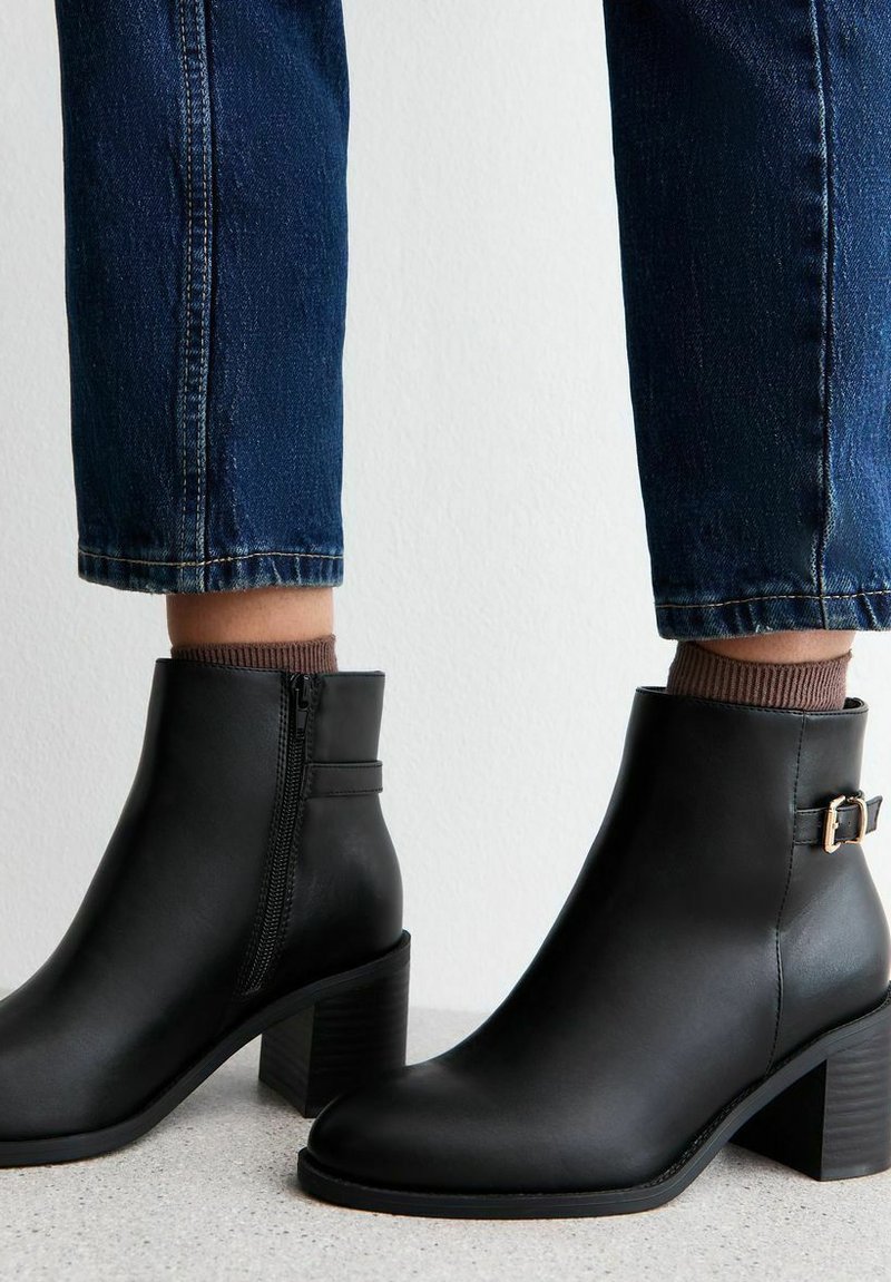 Botas de cuero negro hasta el tobillo con cremallera lateral, puntera cuadrada y tacón cuadrado. Acentuadas con una pequeña hebilla metálica. Llevadas con denim oscuro.