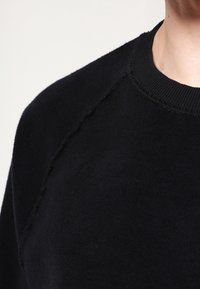 Sudadera negra con una textura suave, cuello redondo y detalles de borde desgastado en los hombros, con un ajuste casual y diseño minimalista.
