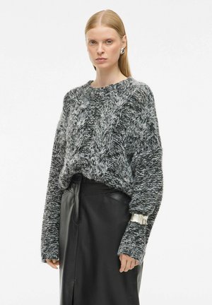 Pull en mailles épaisses noir et gris avec un motif en cable, col rond et coupe ample. Assorti à une jupe en cuir noir ajustée.
