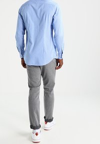 Camisa de botones a rayas azul claro con mangas largas, combinada con pantalones grises y zapatillas blancas. Diseño limpio y moderno con puños enrollados.