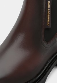 Botas de tornozelo em couro castanho com acabamento brilhante, painéis laterais elásticos e um detalhe com logótipo dourado na parte externa. Sola preta sólida.
