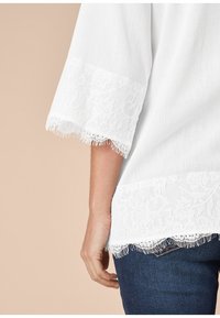 Top blanc avec des manches ¾ bordées de dentelle, présentant de la dentelle florale à l'ourlet et des bords à franges. Porté sur un jean sombre, mettant en valeur le contraste des textures.