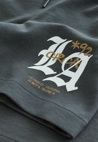 Tissu gris foncé avec texte stylisé "LA" en blanc et or, "1992" et "Los Angeles California 33.987°N 118.4755°W" imprimés sur la surface.