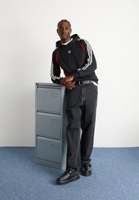Felpa nera con dettagli rossi, jeans grigi a vestibilità ampia e scarpe nere senza lacci con cuciture decorative. In piedi accanto a un armadietto grigio a tre cassetti.