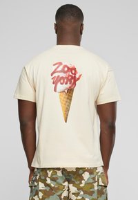 Majica krem boje sa grafikom sladoleda u kornetu sa crvenim sirupom i natpisom "Zoo York" u podebljanom, stiliziranom fontu na leđima.