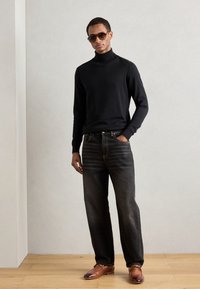 Maglione nero a collo alto, jeans scuri a vestibilità rilassata e scarpe in pelle marrone. Il modello indossa occhiali da sole, posando contro uno sfondo neutro.