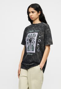 Νεαρή γυναίκα με μακριά σκούρα μαλλιά φοράει μαύρο t-shirt Santa Cruz με φθαρμένο εφέ και μπεζ παντελόνι, στέκεται με το ένα χέρι στην τσέπη.