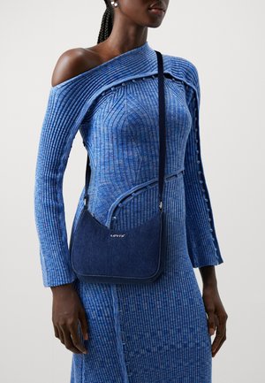 Sac d'épaule en denim bleu à forme incurvée, ferrures argentées, logo embossé et tissu texturé. Porté en bandoulière avec une robe bleue côtelée.