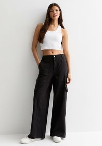 Kvinna med långt hår iklädd en vit kortärmad top, vida svarta cargobyxor, vita sneakers, stående mot en vit vägg med händerna i fickorna.