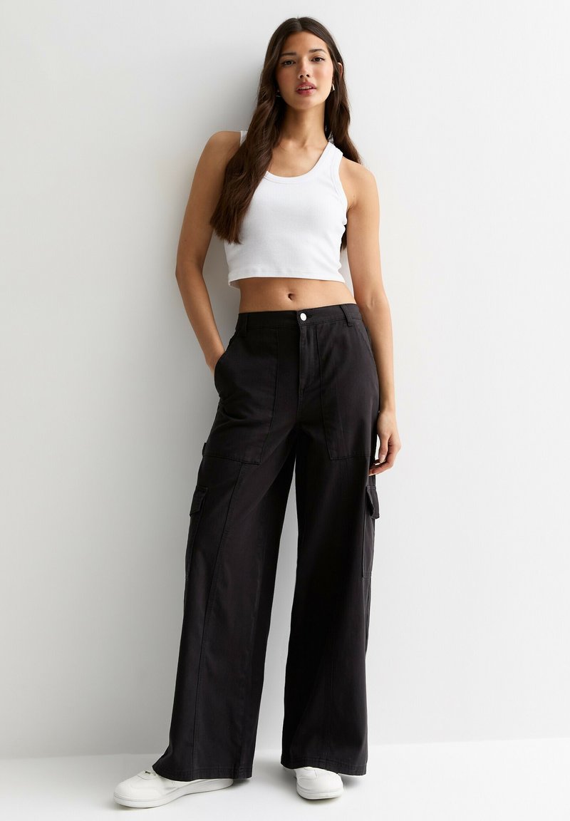 Kvinna med långt hår iklädd en vit kortärmad top, vida svarta cargobyxor, vita sneakers, stående mot en vit vägg med händerna i fickorna.