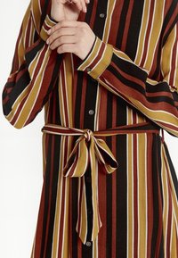 Abito a camicia a righe in rosso, marrone e nero con dettagli bianchi; presenta bottoni e una cintura annodata in vita. Maniche lunghe con polsini a bottone.