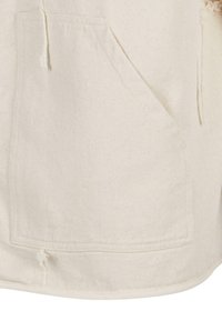 Beige bomullsstoff med en frontlomme, med rå kanter og en teksturert overflate. Slitesterk og uformell design med synlige sømdetaljer.