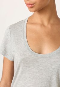 Približnji posnetek osebe, ki nosi svetlo sivo majico z okroglim izrezom in kratkimi rokavi, prikazano je vrat, ramena in zgornji del prsnega koša.