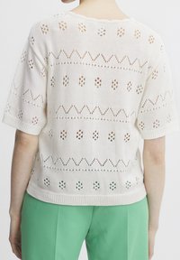 Knit short-sleeve sweater in cream with openwork patterns, featuring zigzag and dot designs. Paired with green trousers.