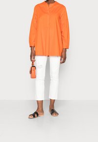 Tunique en coton orange avec un col rond et des manches bouffantes, associée à un jean blanc court et des sandales plates noires. Petit sac à main orange.