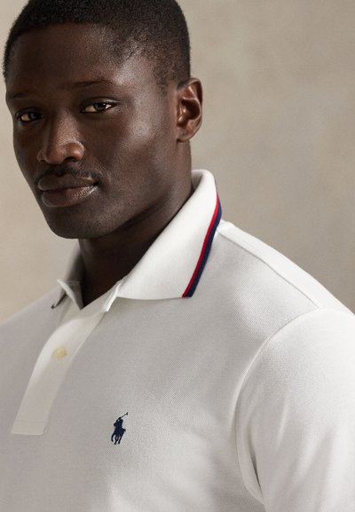 Prim-plan cu un bărbat care poartă un tricou polo alb cu guler tăiat cu albastru închis și roșu și un logo brodat cu un cal în albastru închis.