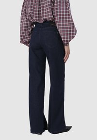 Jean flare en denim bleu foncé à taille haute, avec deux poches arrière et des détails de couture subtils. Porté avec une blouse à carreaux.