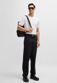 White t-shirt, black wide-leg trousers, black belt, chunky black shoes, and sunglasses. Carrying a black backpack. Neutral background.