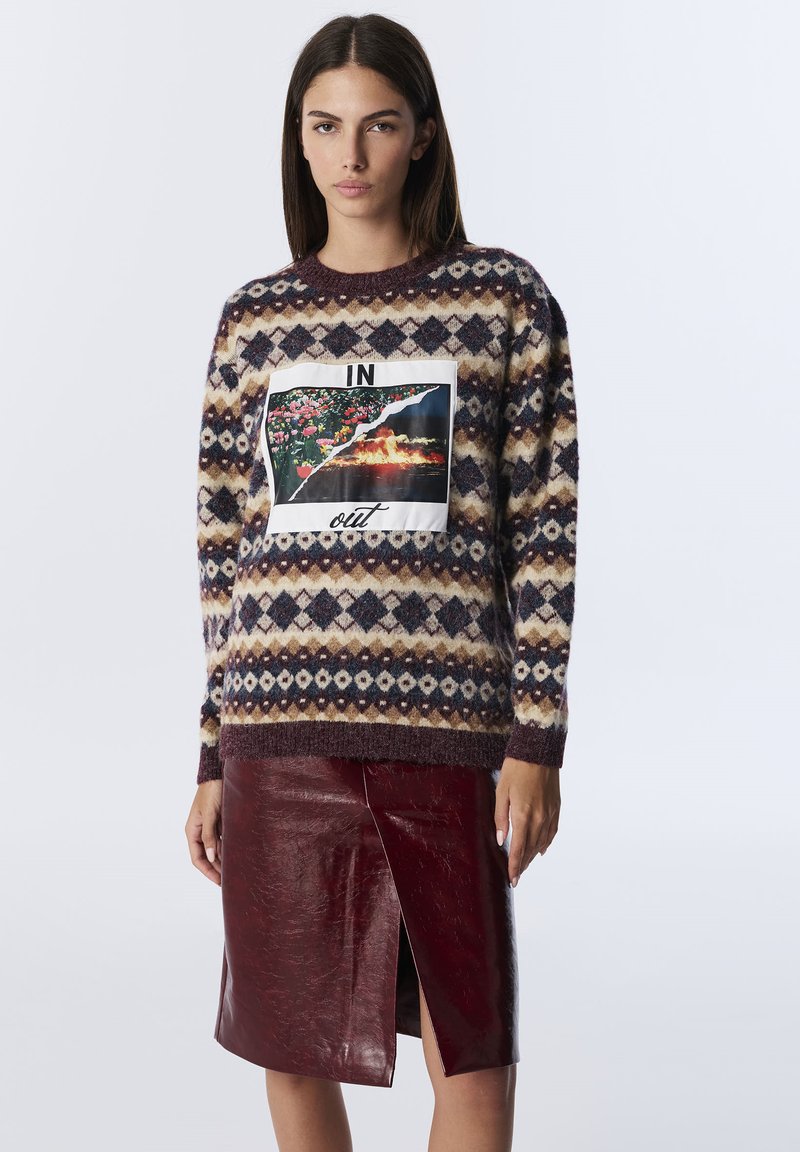 Woman wearing a patterned sweater with a floral and fire graphic labeled "IN" and "out," paired with a shiny burgundy leather skirt with a front slit.