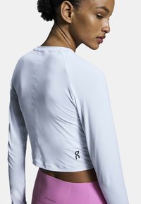 Weißes langärmliges Sportoberteil mit einem cropped Design, aus edlem Stoff und mit einem kleinen schwarzen Logo auf der Rückseite. Kombiniert mit pinken Leggings.