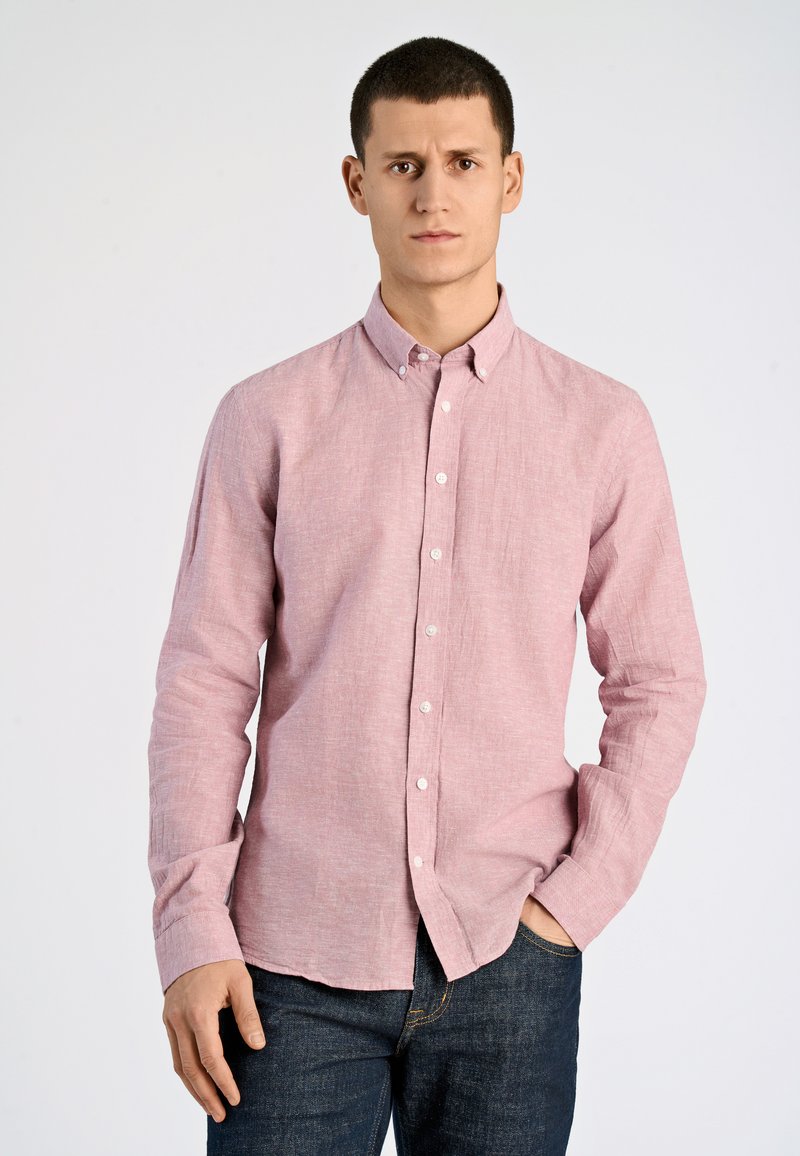 Camicia button-down rosa chiaro con maniche lunghe, realizzata in tessuto texturizzato, abbinata a jeans blu scuro. Colletto strutturato e bottoni frontali.